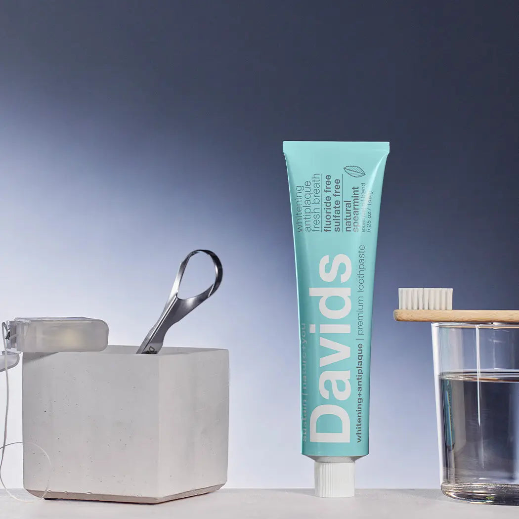 Davids toothpaste tube with a glass of water, wooden toothbrush holder, and metal scoop on a blue gradient background.