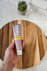 Hand holding a tube of toothpaste on a wooden surface with a plant and glass in the background.